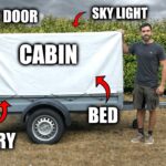 A man building a rustic wooden cabin inside a small trailer with a corrugated plastic roof and acrylic windows.