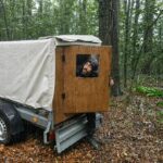 A person stands outside a small wooden cabin built onto a trailer in a wooded area.