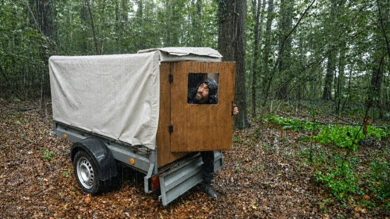 A person stands outside a small wooden cabin built onto a trailer in a wooded area.