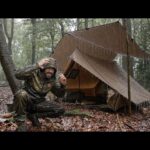 A camper setting up a large tarp over a cabin tent in a rainy forest environment to create a dry workspace.
