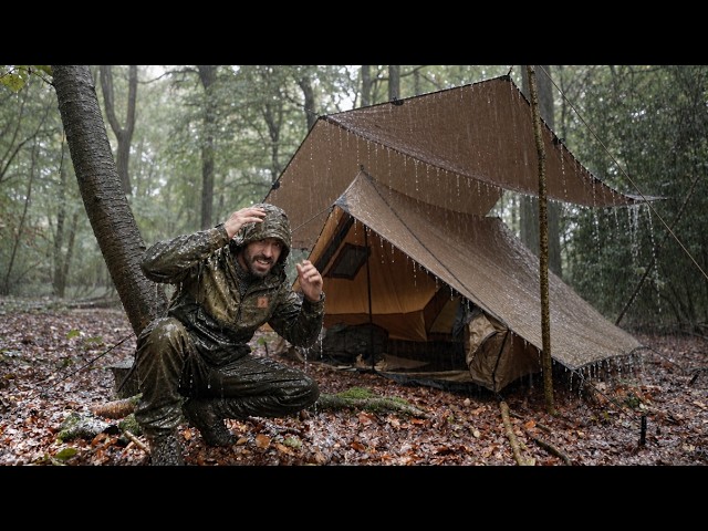 A camper setting up a large tarp over a cabin tent in a rainy forest environment to create a dry workspace.